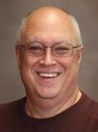 A bald man with glasses is smiling at the camera. He is wearing a dark-colored sweater. The background is plain and neutral.